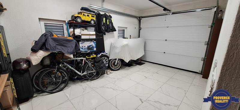 Garáž rodinného domu s bicyklom, policami a bielou keramickou podlahou.